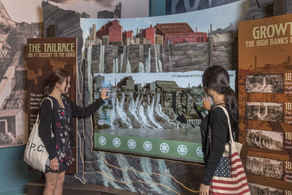 Two women controlling a touchscreen that wipes away an old photo of a mill to reveal a more current photo