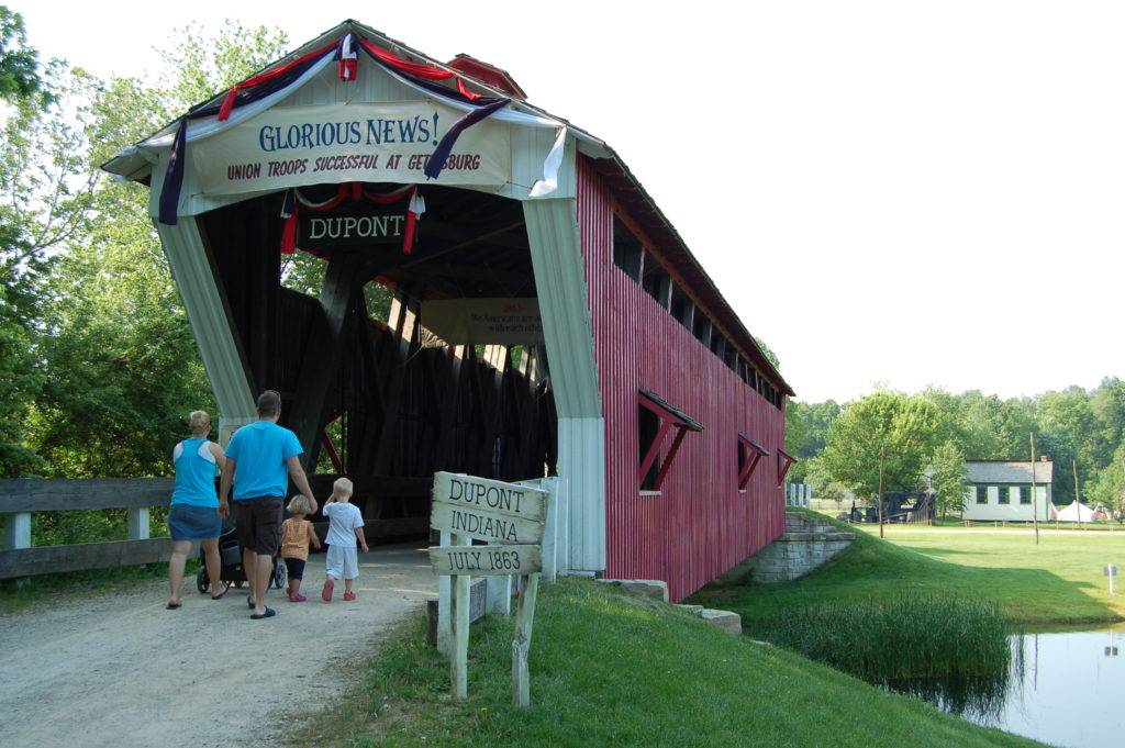 Family entering a red covered bridge in Dupont Indiana. A banner reads Glorious News! Union Troops Successful at Gettysburg