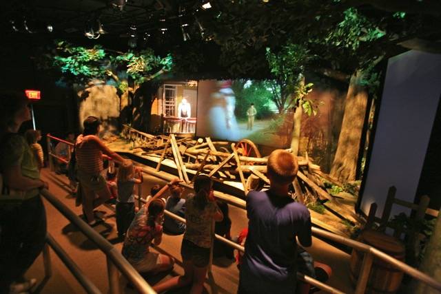 Family watching an immersive theater from behind the rails. The theater contains fake trees, logs, a wagon wheel, a boy on a balcony, and a projection screen