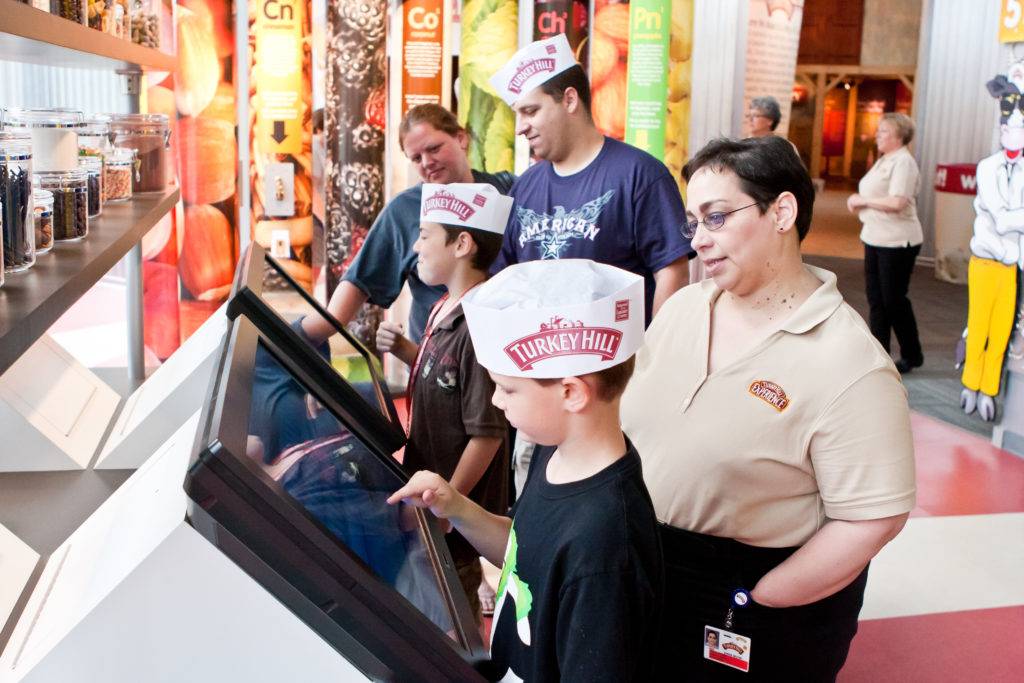 Children wearing Turkey Hill hats and using a touch screen interactive, while assisted by parent's and exhibit staff